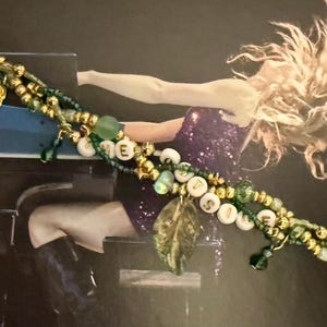 K&ouml;nnte beinhalten: Ein mehrstr&auml;ngiges Armband mit gr&uuml;nen und goldenen Perlen, Blattanh&auml;ngern und wei&szlig;en Buchstabenperlen, die ein Wort buchstabieren. Das Armband ist vor dem Hintergrund einer Person, die Klavier spielt, platziert.