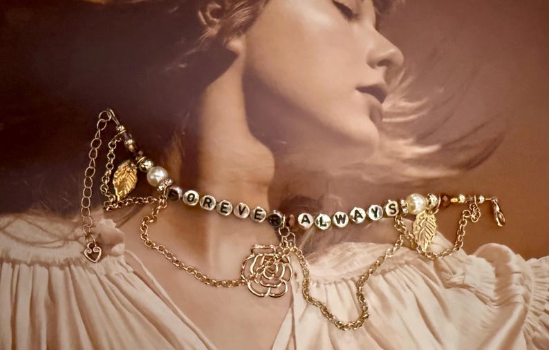 Op de afbeelding: Goudkleurige ketting met de woorden "FOREVER ALWAYS" gespeld in kleine, ronde, witte letterkralen. De ketting heeft een rozenhanger, bladbedels en parelaccenten. De achtergrond is een zachte, vage afbeelding.