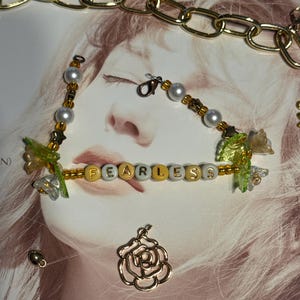 Op de afbeelding: Een kralenarmband met het woord "FEARLESS" in gouden en witte letterkralen. De armband heeft parels, gouden accenten en decoratieve bloemelementen. Een gouden ketting en een roosvormige bedel zijn ook zichtbaar.