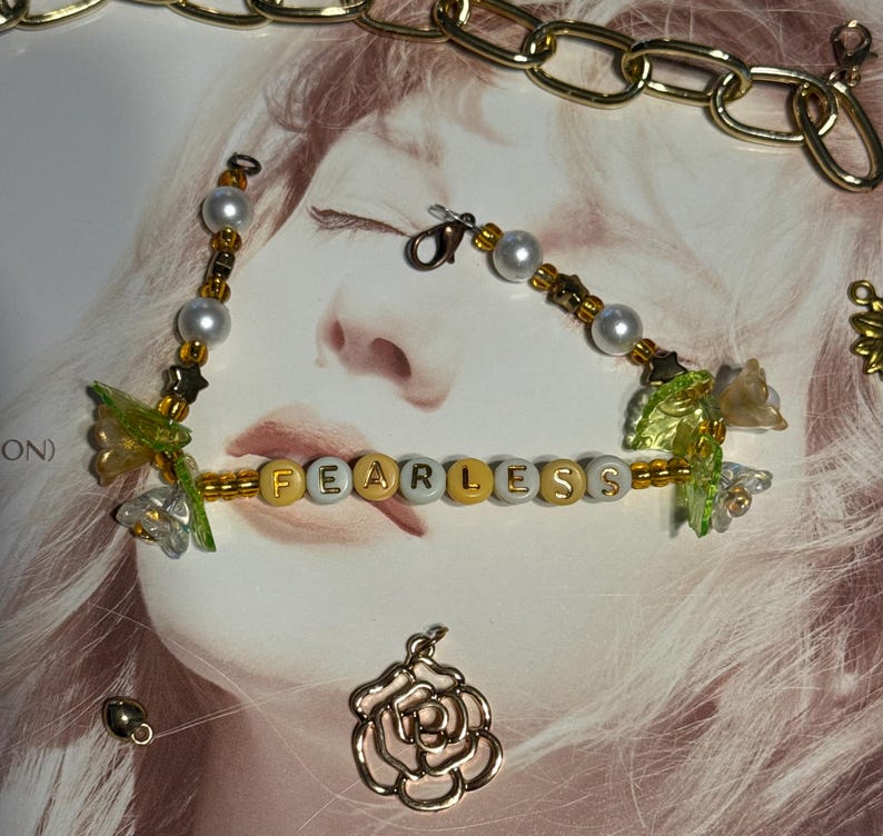 Op de afbeelding: Een kralenarmband met het woord "FEARLESS" in gouden en witte letterkralen. De armband heeft parels, gouden accenten en decoratieve bloemelementen. Een gouden ketting en een roosvormige bedel zijn ook zichtbaar.
