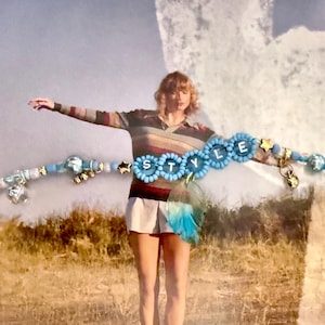 May include: A beaded bracelet with the word "STYLE" spelled out in blue flower-shaped beads. The bracelet features various blue, silver, and gold beads, charms, and a chain. The background shows a person in a striped shirt and shorts.