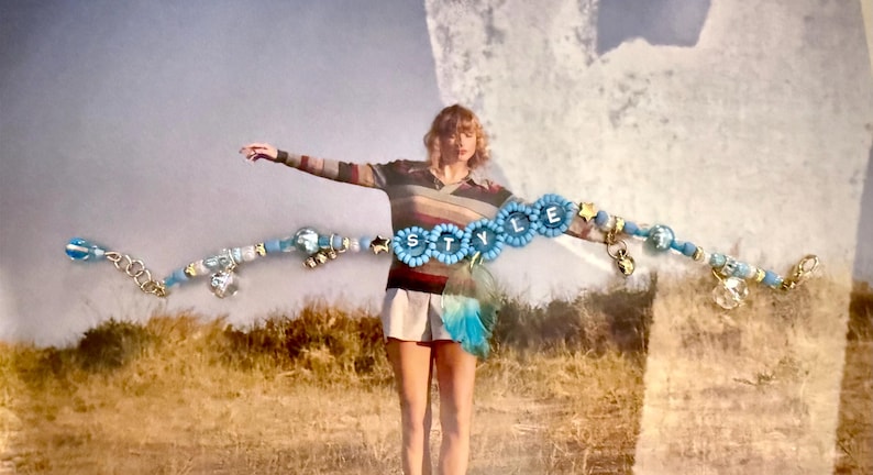 May include: A beaded bracelet with the word "STYLE" spelled out in blue flower-shaped beads. The bracelet features various blue, silver, and gold beads, charms, and a chain. The background shows a person in a striped shirt and shorts.
