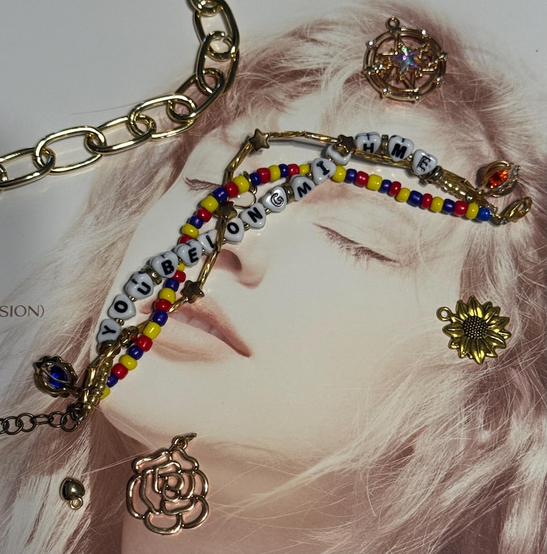 Op de afbeelding: Goudkleurige sieraden, waaronder een ketting, kralenarmbanden met de woorden "YOU BELONG TO ME" en diverse bedels zoals een zonnebloem en een roos. De armbanden hebben rode, gele en blauwe kralen.