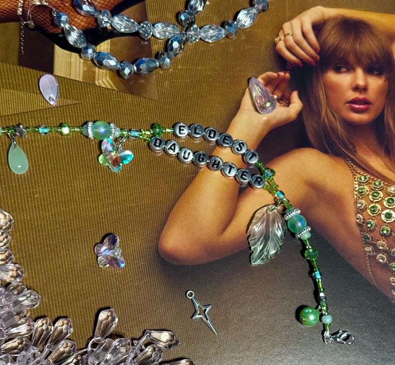 Puede incluir: Una pulsera de cuentas con las palabras "ELDEST DAUGHTER" escritas con cuentas de letras plateadas. La pulsera presenta cuentas verdes y transparentes, dijes de mariposa y un dije de hoja. Tambi&eacute;n se ven joyas de cuentas adicionales.