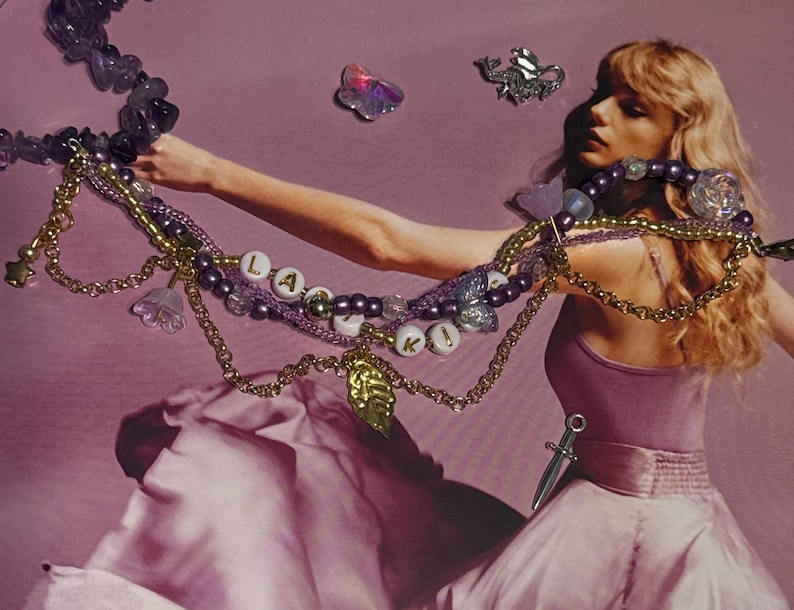 Puede incluir: Una colecci&oacute;n de collares y cadenas de cuentas en tonos morados y dorados. Un collar deletrea la palabra "L A K I" con cuentas de letras blancas. La imagen tiene una est&eacute;tica suave y so&ntilde;adora, con una mujer con un vestido rosa en el fondo.