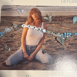 May include: A beaded bracelet with the word "1989" spelled out in white letter beads. The bracelet features blue and white beads, along with charms including a paper airplane, a heart, and a dove. The bracelet is on a beach background.