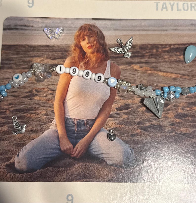 May include: A beaded bracelet with the word "1989" spelled out in white letter beads. The bracelet features blue and white beads, along with charms including a paper airplane, a heart, and a dove. The bracelet is on a beach background.
