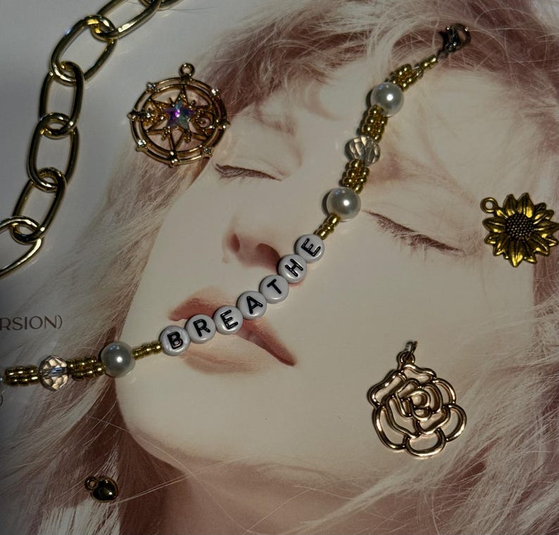 Op de afbeelding: Een goudkleurige ketting en diverse bedels op een lichte achtergrond. De bedels omvatten een kompas, een zonnebloem en een roos. Een kralenarmband spelt het woord "breathe" met witte letterkralen en parelaccenten.