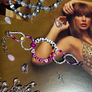 Puede incluir: Pulseras de cuentas con las palabras "BORING BARBIE" deletreadas con cuentas de letras blancas y rosas. Las pulseras est&aacute;n acentuadas con cuentas rosas y moradas y una cadena plateada. Joyas adicionales y elementos decorativos est&aacute;n dispersos.