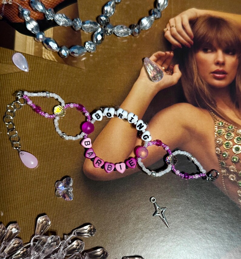 Puede incluir: Pulseras de cuentas con las palabras "BORING BARBIE" deletreadas con cuentas de letras blancas y rosas. Las pulseras est&aacute;n acentuadas con cuentas rosas y moradas y una cadena plateada. Joyas adicionales y elementos decorativos est&aacute;n dispersos.