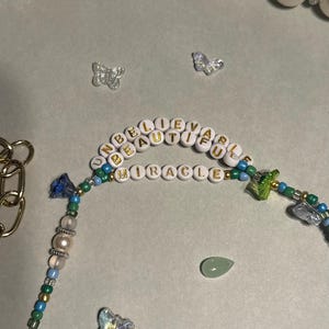 K&ouml;nnte beinhalten: Eine Perlenkette mit den Worten "UNBELIEVABLE BEAUTIFUL MIRACLE" in goldenen Buchstaben. Die Kette hat blaue, gr&uuml;ne und goldene Perlen, einen gr&uuml;nen Tropfenstein und Schmetterlingsanh&auml;nger.
