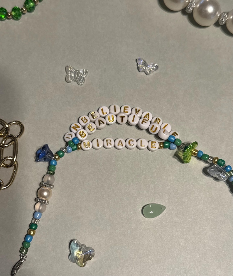 K&ouml;nnte beinhalten: Eine Perlenkette mit den Worten "UNBELIEVABLE BEAUTIFUL MIRACLE" in goldenen Buchstaben. Die Kette hat blaue, gr&uuml;ne und goldene Perlen, einen gr&uuml;nen Tropfenstein und Schmetterlingsanh&auml;nger.