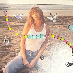May include: A colorful beaded bracelet with the word "SLUT" spelled out in turquoise beads. The bracelet features a mix of orange, pink, yellow, and turquoise beads. The background includes a person wearing a white tank top and blue jeans.
