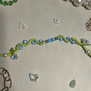 K&ouml;nnte beinhalten: Ein Perlenarmband mit den blauen Buchstaben "COLD AS YOU", abwechselnd mit gr&uuml;nen und blauen Perlen. Das Bild zeigt auch eine Perlenkette, eine goldene Kette und kleine Schmetterlingsanh&auml;nger, alles auf einer hellgrauen Oberfl&auml;che.