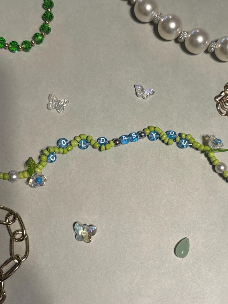 K&ouml;nnte beinhalten: Ein Perlenarmband mit den blauen Buchstaben "COLD AS YOU", abwechselnd mit gr&uuml;nen und blauen Perlen. Das Bild zeigt auch eine Perlenkette, eine goldene Kette und kleine Schmetterlingsanh&auml;nger, alles auf einer hellgrauen Oberfl&auml;che.