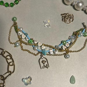 K&ouml;nnte beinhalten: Eine handgefertigte Halskette mit goldenen Ketten, wei&szlig;en und blauen Perlen und Buchstabenperlen mit der Aufschrift "LOVE". Die Halskette hat einen Rosenanh&auml;nger und ist von anderen Schmuckelementen umgeben, darunter ein goldenes Kettenarmband und Perlenketten.
