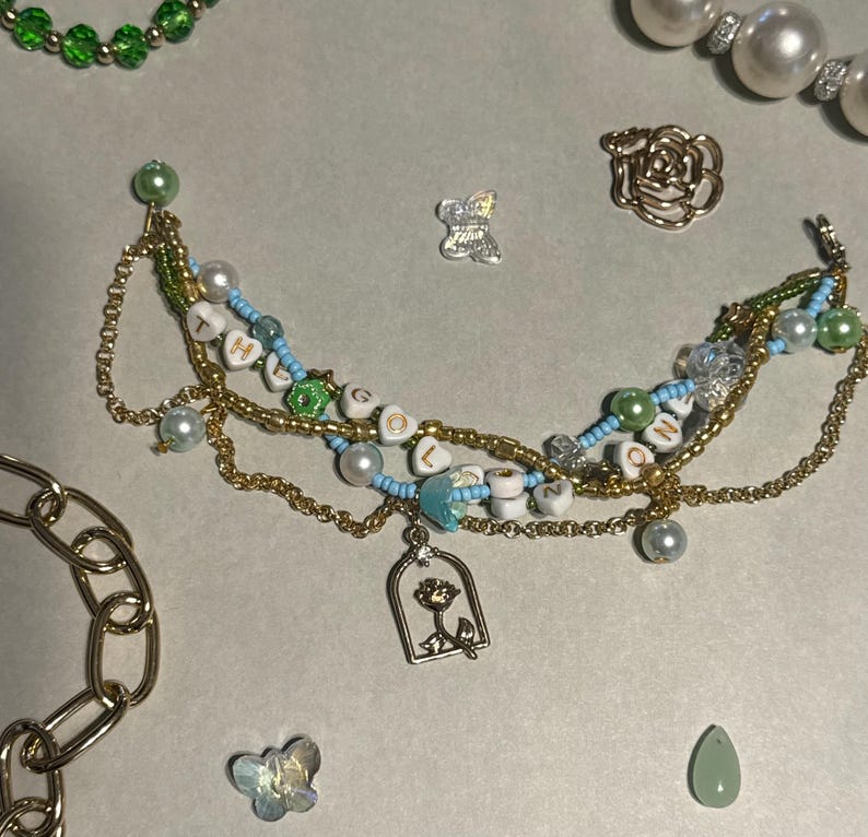K&ouml;nnte beinhalten: Eine handgefertigte Halskette mit goldenen Ketten, wei&szlig;en und blauen Perlen und Buchstabenperlen mit der Aufschrift "LOVE". Die Halskette hat einen Rosenanh&auml;nger und ist von anderen Schmuckelementen umgeben, darunter ein goldenes Kettenarmband und Perlenketten.