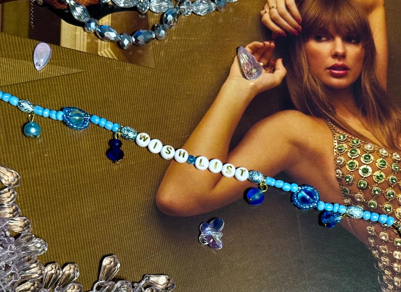 Puede incluir: Una pulsera de cuentas azules con la palabra "WISH LIST" escrita con cuentas de letras blancas. La pulsera tiene abalorios azules y est&aacute; colocada sobre un fondo dorado. Tambi&eacute;n se ven otras joyas.