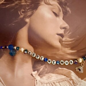 Op de afbeelding: Een armband met de woorden "TELL ME WHY" gespeld in witte letterkralen. De armband heeft blauwe en gouden kralen, met een bloemvormige bedel. De achtergrond is een sepia-kleurige afbeelding van een persoon.