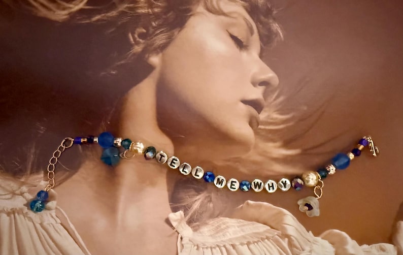 Op de afbeelding: Een armband met de woorden "TELL ME WHY" gespeld in witte letterkralen. De armband heeft blauwe en gouden kralen, met een bloemvormige bedel. De achtergrond is een sepia-kleurige afbeelding van een persoon.