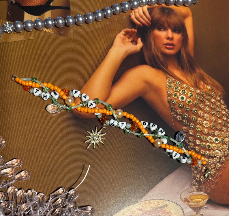 Puede incluir: Una pulsera de cuentas con las palabras "THE FAT OF HOPE" deletreadas con cuentas de letras blancas. La pulsera presenta cuentas naranjas y verdes, un dije de estrella y un dije transparente. Se ve una segunda hebra de cuentas grises.