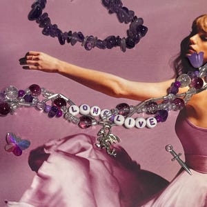 Puede incluir: Un collar con las palabras "LONG LIVE" en cuentas de letras blancas, acentuado con cuentas moradas y un dije de drag&oacute;n plateado. Un segundo collar de piedras moradas est&aacute; arriba. Una peque&ntilde;a daga plateada y una mariposa tambi&eacute;n est&aacute;n en la imagen.