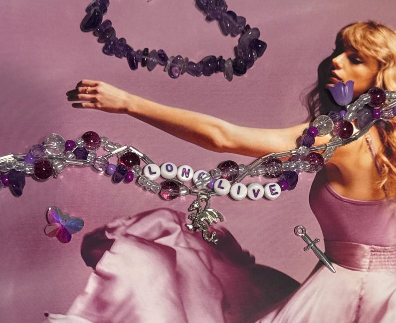 Puede incluir: Un collar con las palabras "LONG LIVE" en cuentas de letras blancas, acentuado con cuentas moradas y un dije de drag&oacute;n plateado. Un segundo collar de piedras moradas est&aacute; arriba. Una peque&ntilde;a daga plateada y una mariposa tambi&eacute;n est&aacute;n en la imagen.