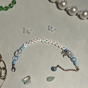 K&ouml;nnte beinhalten: Ein Armband mit Perlen, auf dem der Name "MARYSONG" in wei&szlig;en Buchstabenperlen steht. Das Armband hat blaue und perlmuttfarbene Perlen, Schmetterlingsanh&auml;nger und einen goldfarbenen Schleifenanh&auml;nger. Weitere Schmuckst&uuml;cke sind eine Perlenkette und eine goldene Kette.