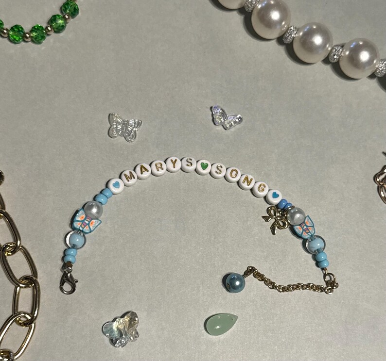 K&ouml;nnte beinhalten: Ein Armband mit Perlen, auf dem der Name "MARYSONG" in wei&szlig;en Buchstabenperlen steht. Das Armband hat blaue und perlmuttfarbene Perlen, Schmetterlingsanh&auml;nger und einen goldfarbenen Schleifenanh&auml;nger. Weitere Schmuckst&uuml;cke sind eine Perlenkette und eine goldene Kette.