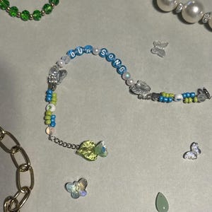 K&ouml;nnte beinhalten: Ein Armband mit Perlen, auf dem die Worte "OUR SONG" in blauen Perlen stehen. Das Armband hat eine Mischung aus blauen, gr&uuml;nen und wei&szlig;en Perlen, Schmetterlingsanh&auml;ngern und einem gr&uuml;nen Edelstein. Weitere Schmuckst&uuml;cke sind ebenfalls zu sehen.
