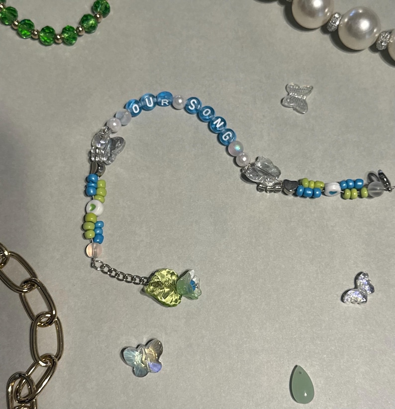 K&ouml;nnte beinhalten: Ein Armband mit Perlen, auf dem die Worte "OUR SONG" in blauen Perlen stehen. Das Armband hat eine Mischung aus blauen, gr&uuml;nen und wei&szlig;en Perlen, Schmetterlingsanh&auml;ngern und einem gr&uuml;nen Edelstein. Weitere Schmuckst&uuml;cke sind ebenfalls zu sehen.