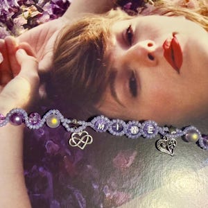 Puede incluir: Una pulsera de cuentas moradas con la palabra "MINE" escrita con cuentas blancas. La pulsera incluye dijes en forma de coraz&oacute;n y cuentas decorativas. Tambi&eacute;n es visible un dije en forma de estrella.