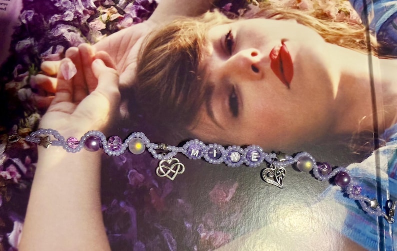 Puede incluir: Una pulsera de cuentas moradas con la palabra "MINE" escrita con cuentas blancas. La pulsera incluye dijes en forma de coraz&oacute;n y cuentas decorativas. Tambi&eacute;n es visible un dije en forma de estrella.