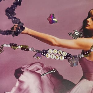 Puede incluir: Un collar con las palabras "LONG LIVE" deletreadas en cuentas de letras blancas. El collar presenta cuentas moradas, dijes de flores y dijes de mariposas. Tambi&eacute;n es visible un segundo collar con cuentas de piedra morada.