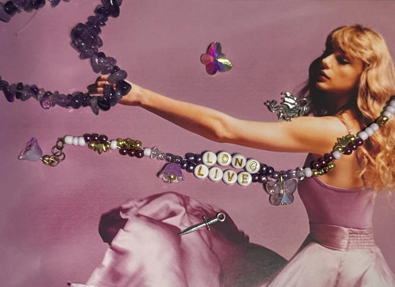 Puede incluir: Un collar con las palabras "LONG LIVE" deletreadas en cuentas de letras blancas. El collar presenta cuentas moradas, dijes de flores y dijes de mariposas. Tambi&eacute;n es visible un segundo collar con cuentas de piedra morada.