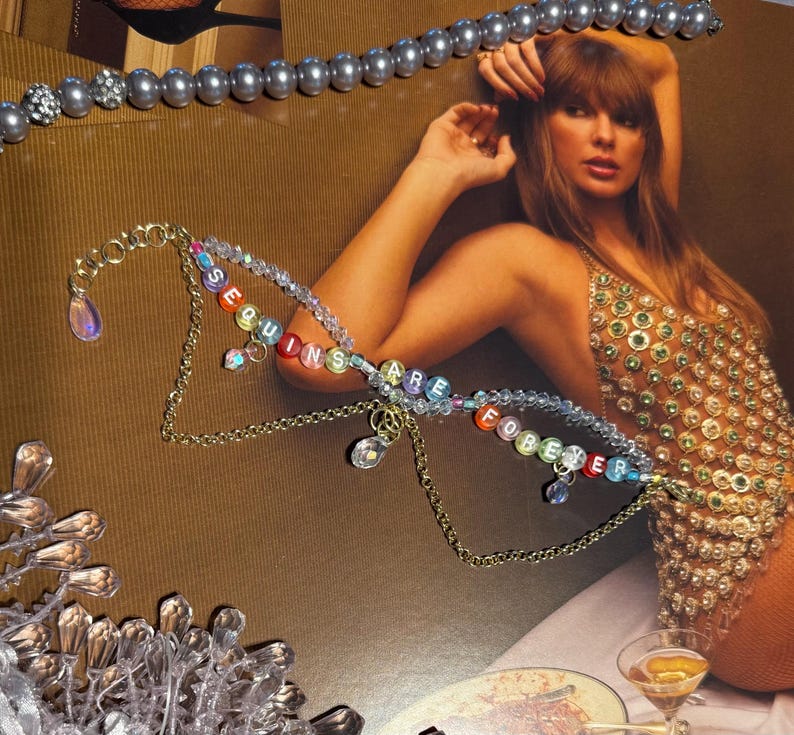 Puede incluir: Un collar dorado con las palabras "SEQUINS ARE FOREVER" en cuentas de letras coloridas. El collar tiene colgantes. Una collar de perlas est&aacute; en la parte superior de la imagen. El fondo es de color marr&oacute;n.