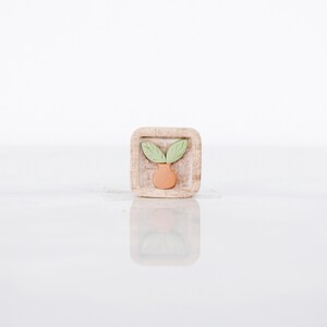 May include: A small, square wooden block with a carved design of a terracotta-colored vase with two green leaves. The block has a natural wood grain finish and is reflected in the glossy surface below.
