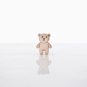 May include: A small, light brown teddy bear figurine with a simple design. The bear has a rounded body, small arms and legs, and a detailed face with dark brown eyes and a nose. The figurine is standing on a reflective surface.