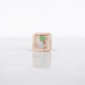 May include: A small, square wooden tray with a carved design of a bulbous vegetable with green leaves. The vegetable is light gray with a textured surface. The leaves are a bright green color.