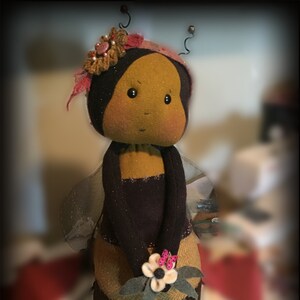 May include: A handmade felt bee doll with black and yellow stripes, wearing a pink and brown flower crown and holding a white flower with pink accents.