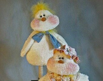 May include: Two white plush toy chickens with pink cheeks and yellow beaks. The chicken on the left has a blue bow tie and the chicken on the right has a pink and yellow bow. Both chickens have long legs and are standing on a wooden box.