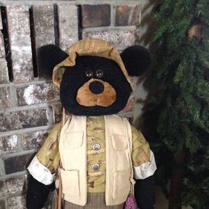 May include: A black bear plush toy dressed in a fishing outfit. The bear is wearing a brown and white plaid shirt, a tan vest with buttons, and brown pants. The bear is holding a fishing rod and has a string of purple and pink fish hanging from its hand.