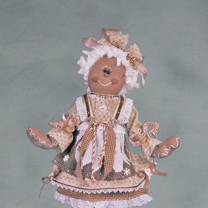 May include: A gingerbread doll with brown hair, wearing a brown and white dress with a brown and white checkered apron, and brown shoes with red accents. The doll is standing on a wooden base with white frosting.
