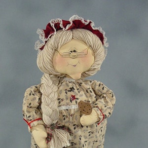 May include: A handmade doll with a white braid, wearing a red and white dress with a floral pattern, holding a brown cookie. The doll is wearing red shoes with green accents.