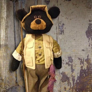 May include: A plush black bear dressed in a brown and beige fishing vest, holding a fishing rod and a purple fish. The bear is standing on a wooden base.