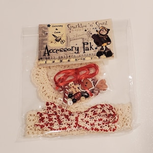 Puede incluir: Una bolsa de plástico transparente contiene suministros para manualidades. En el interior hay un ribete de encaje color crema, una cinta roja y pequeños adornos, incluyendo hombres de jengibre y botones. El paquete está etiquetado como "Accessory Pak".