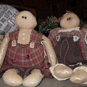 Peut inclure: Deux poupées en pain d'épices faites à la main habillées de tissu à carreaux rouge et brun. Les poupées ont des coutures blanches autour de leurs bords et des yeux noirs.
