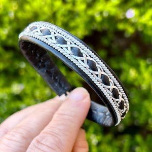 May include: A black leather bracelet with a silver braided design. The bracelet is a simple, elegant design.