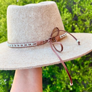 May include: A beige fedora-style hat with a wide brim and a brown leather band with a decorative pattern. The hat has a matching brown leather tie. The hat is held up against a green background.
