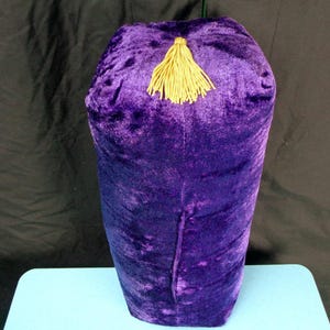 May include: A cylindrical, purple velvet object with a gold tassel. The velvet has a plush texture. The object is likely a decorative item or container. The background is dark. The item is approximately 30 cm tall.